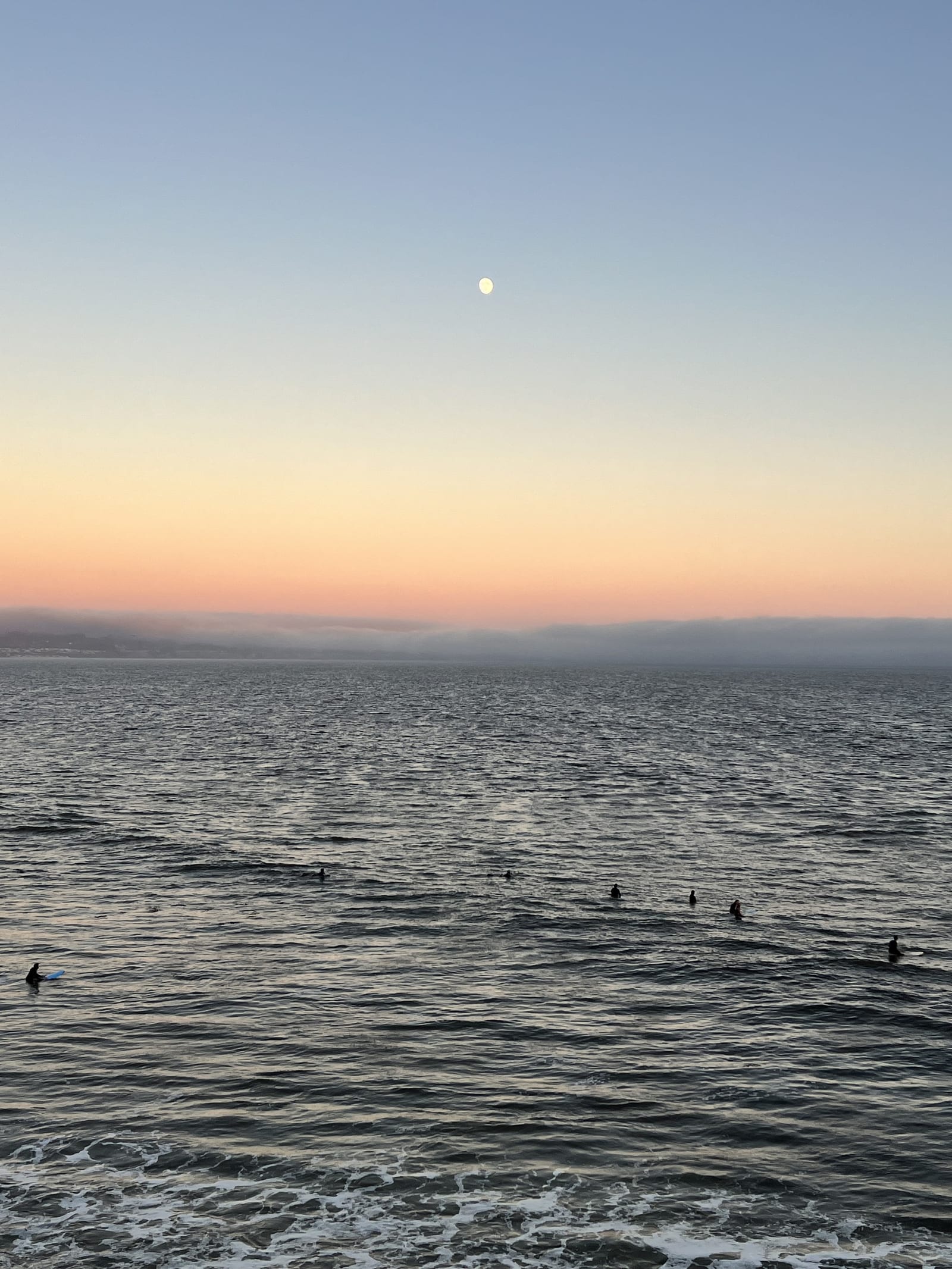 The moon rising over the ocean at dusk off Santa Cruz, with surfers waiting for waves
