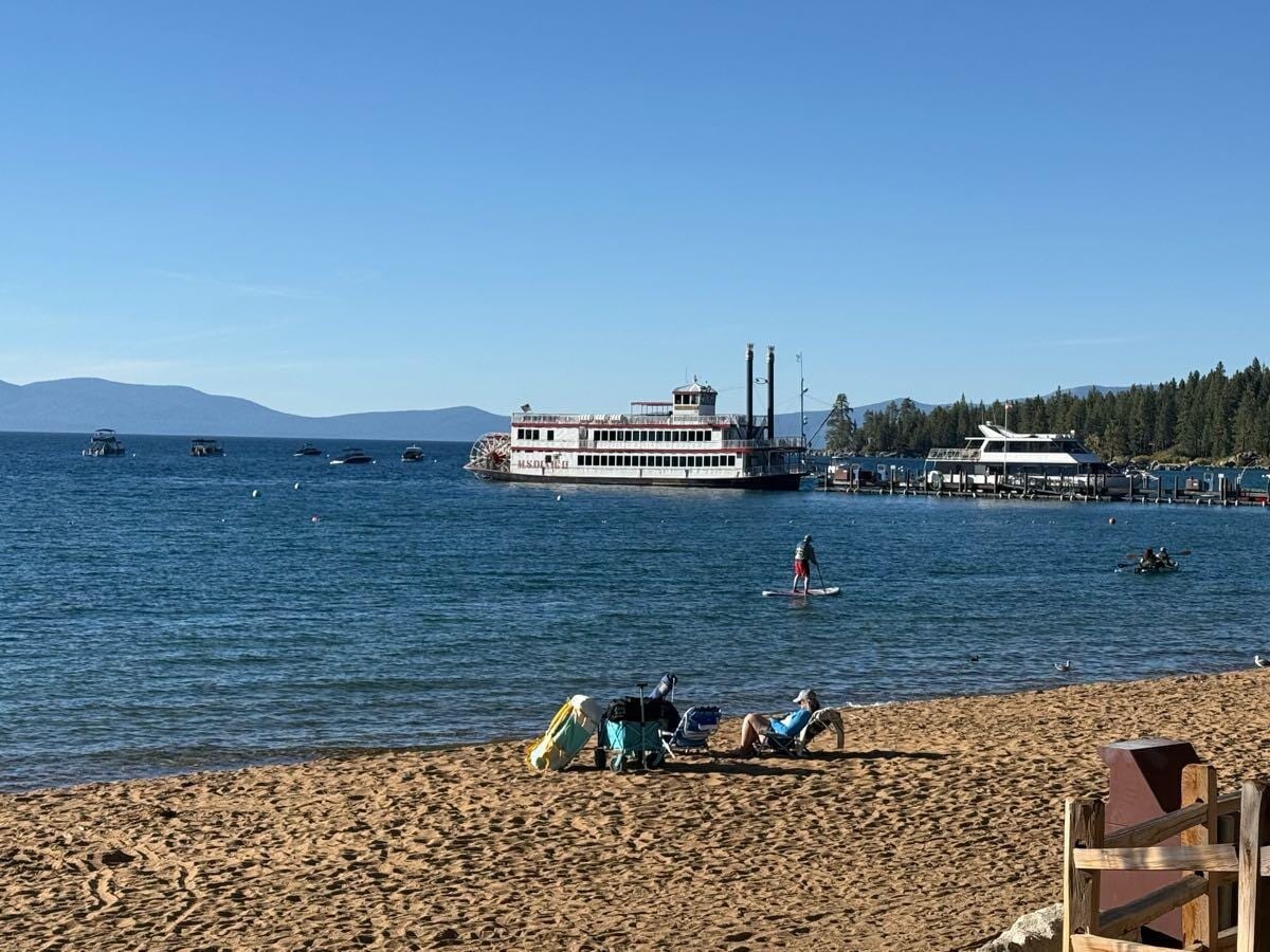 Sandy Lake Tahoe beach on a clear day with a paddlewheel boat, a paddleboarder and distant mountains