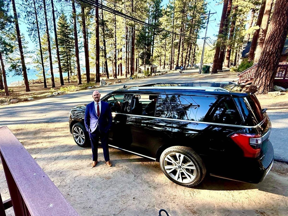 Jon in a navy suit standing beside his black SUV among tall pines at Lake Tahoe