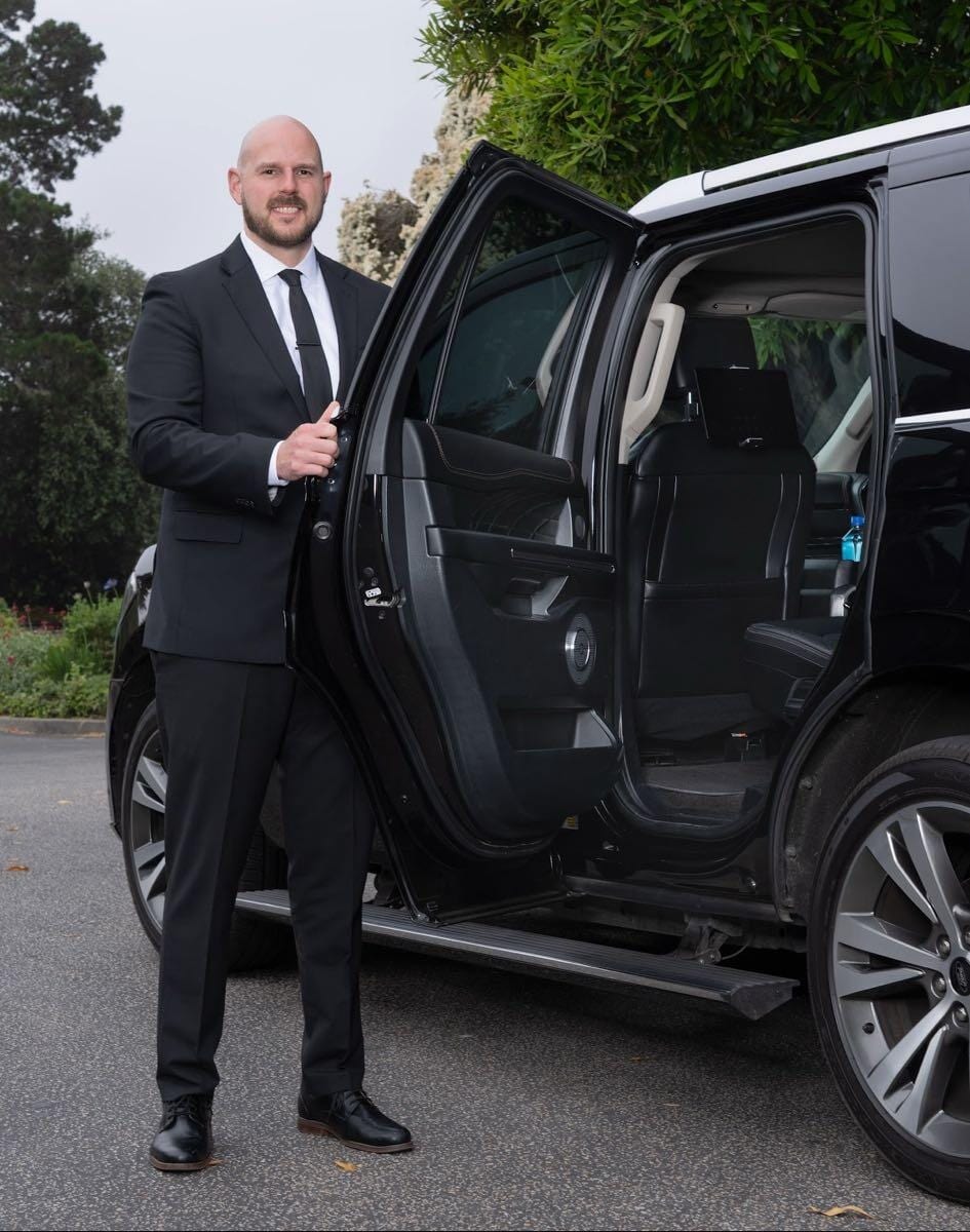 Jon in a black suit and tie holding open the rear door of his black SUV